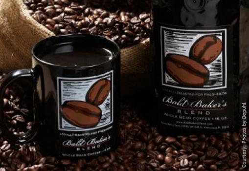 Photo of a Bald Baker's Blend coffee mug displaying a cartoon image of two beans. Cup is full of dark coffee. to the right in the background is a bag of coffee with the label the same as the design on the mug. More loose coffee beans are in the background and foreground.