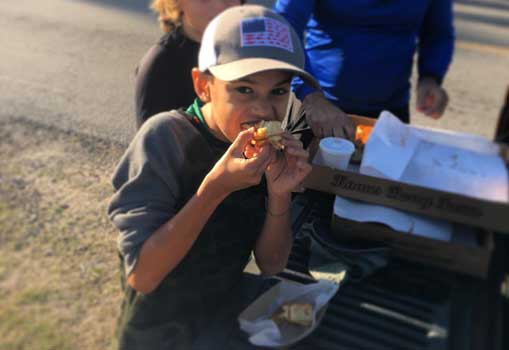 Photo of a boy about 8 years old wearing a baseball cap with an American Flag on it and he is eating a cinnamon bun. in the background are two boxes of buns on the tailgate of a truck.