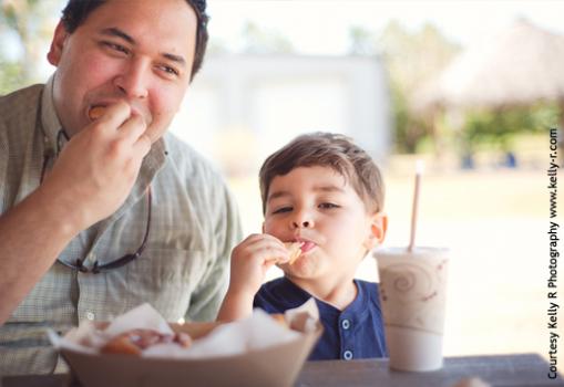 Photo of a father and very young son enjoying cinnamon rolls, with a basket of rolls and a milkshake in the foreground and the farm tiki hut in the background (blurry).